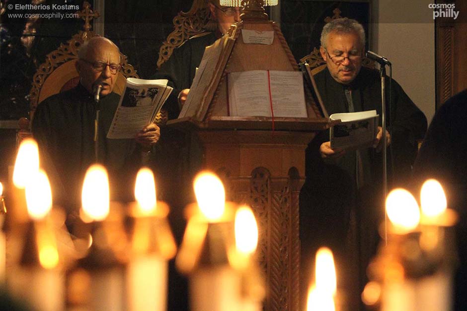 Two chanters read from liturgical books at the psalterion during a Greek Orthodox church service, with candlelight in the foreground.