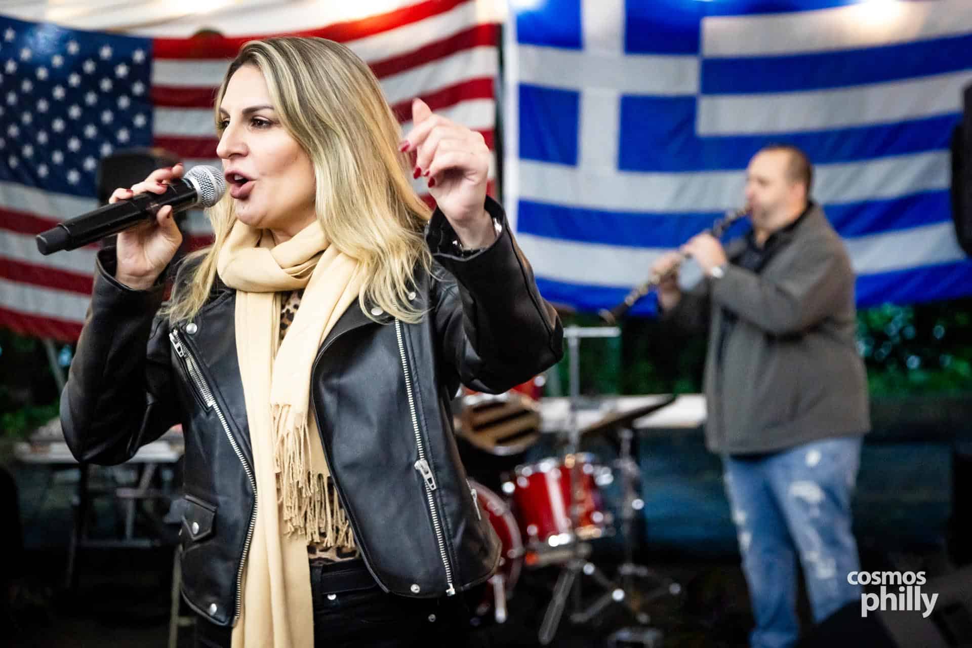 Singer performs at the OPA Greek Festival at Annunciation Church in Elkins Park with Greek and American flags in the background