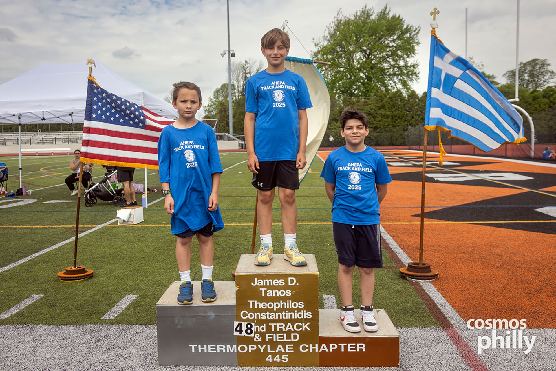 Youth participants stand on the awards podium at the 2025 AHEPA Pan-Hellenic Track and Field Meet, with American and Greek flags in the background.