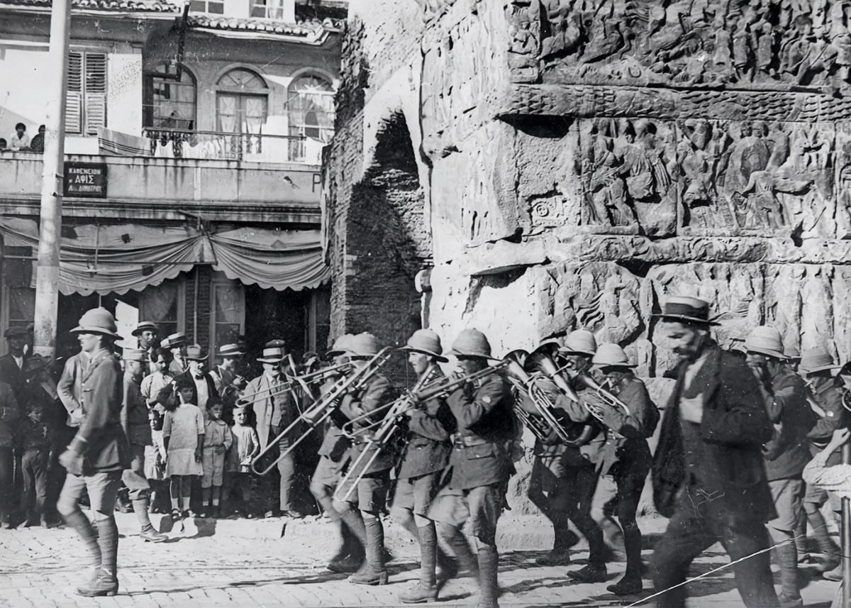 In August 1916, during World War I, an Italian military band parades past the Arch of Galerius in Thessaloniki, while local residents watch in silence.