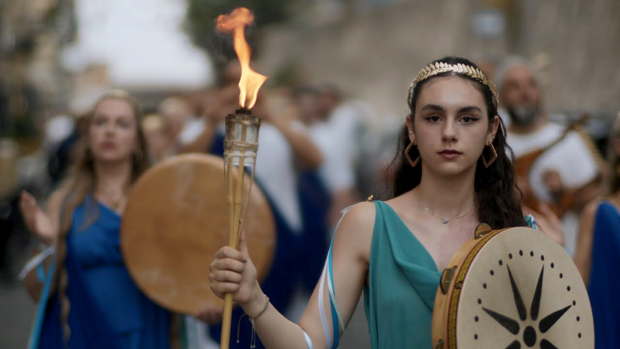 Young woman in ancient Greek costume holding a flaming torch and drum during the torchlight procession in Naples, part of the Magna Graecia Fest honoring Parthenope