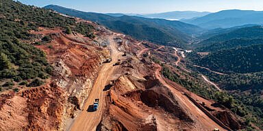 Aerial view of a bauxite mining site in Central Greece with red soil, terraced excavation, and trucks operating in a mountainous quarry under a clear blue sky