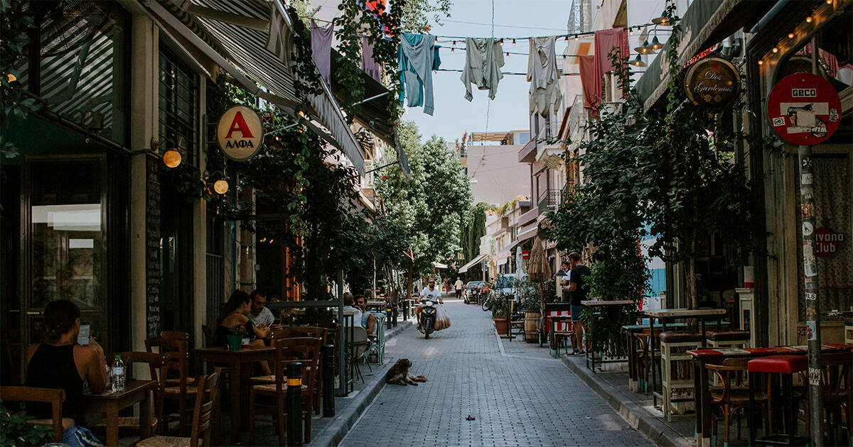 Bustling narrow street in Athens with outdoor cafés, laundry hanging above, and people enjoying a summer afternoon.