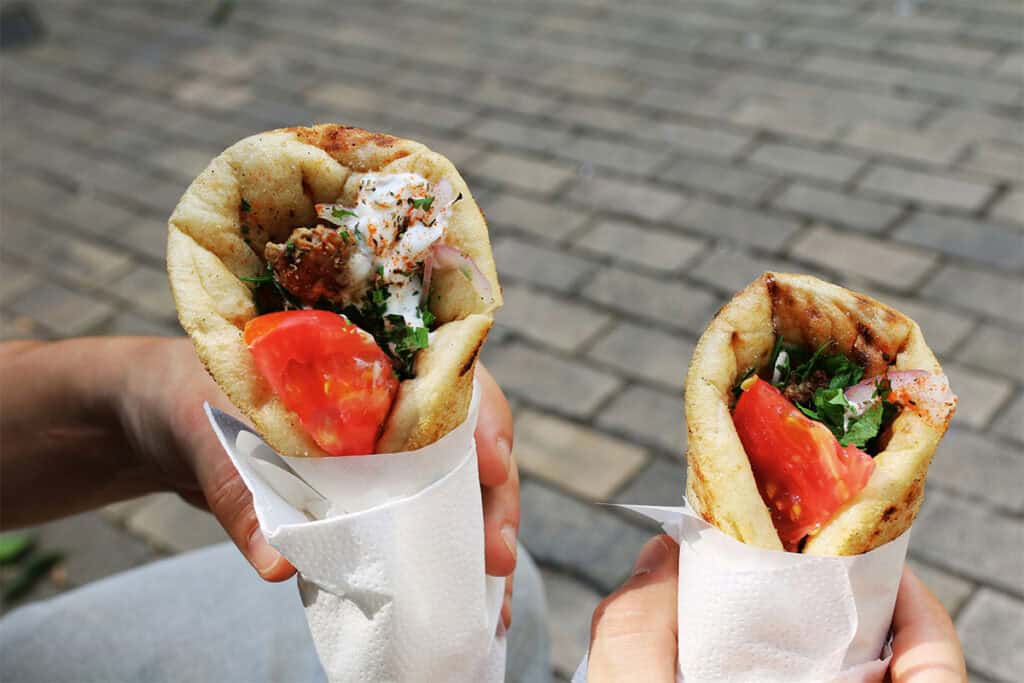 Two smaller gyros held in hand, with tomato, herbs, and light toppings, served in grilled pita on a street in Athens