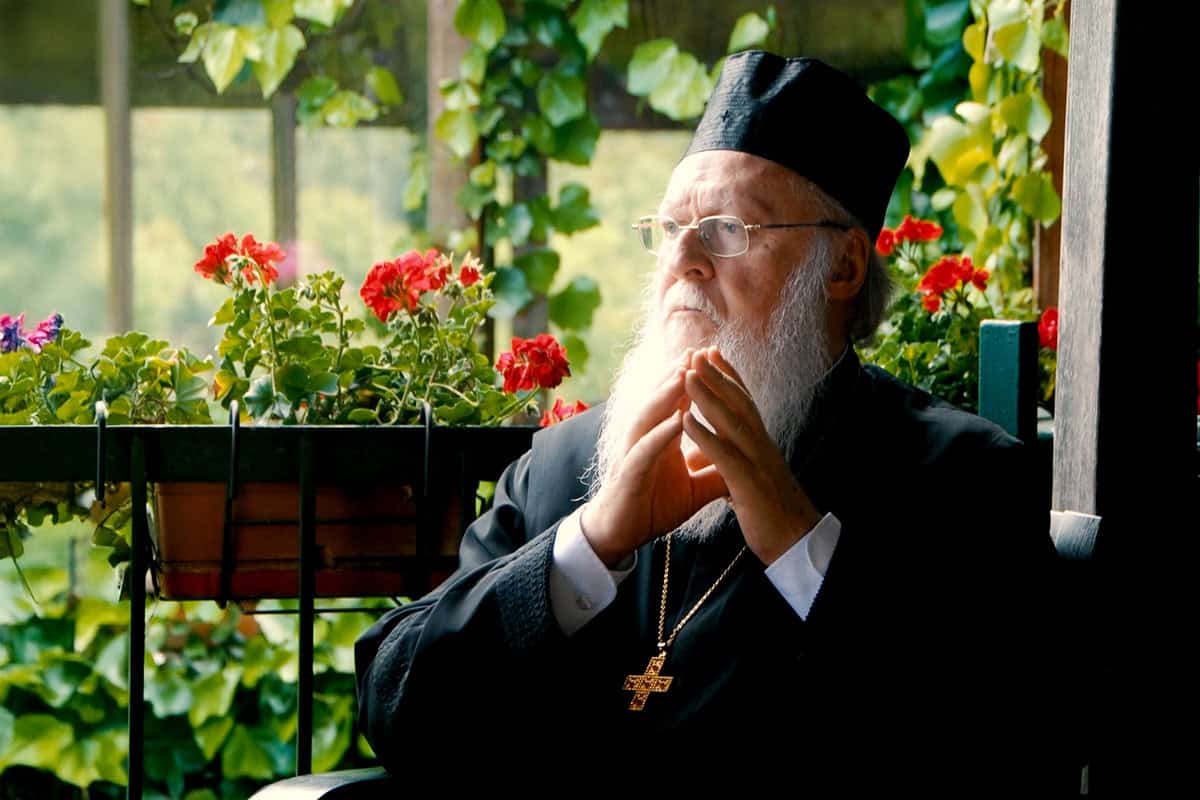 Ecumenical Patriarch Bartholomew in a contemplative moment, seated outdoors with flowers in the background