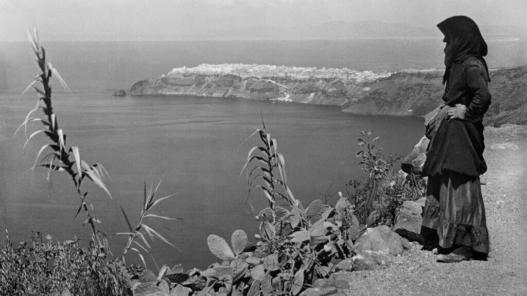 Woman in traditional dress standing above the Santorini caldera, facing a village across the bay.