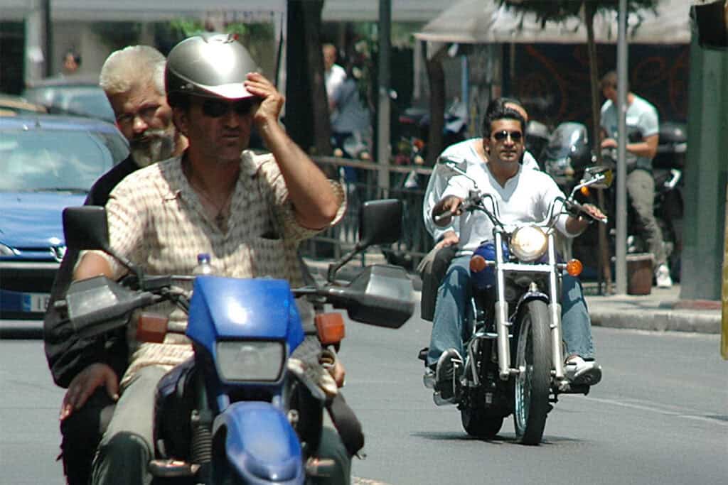 Motorcyclists riding in Greece, one adjusting his helmet while others ride without