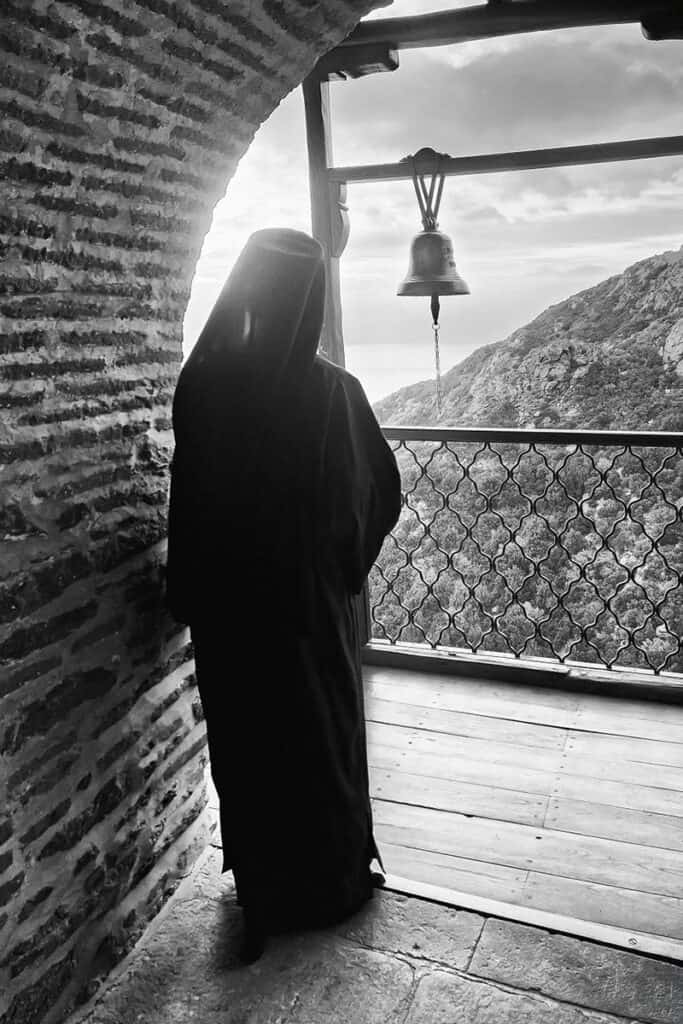 Monk standing in an archway beside a bell, looking out over the mountains at Mount Athos.