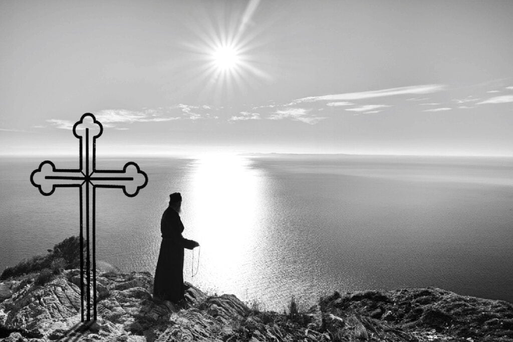 Monk standing with prayer rope beside a cross at sunset on Mount Athos.