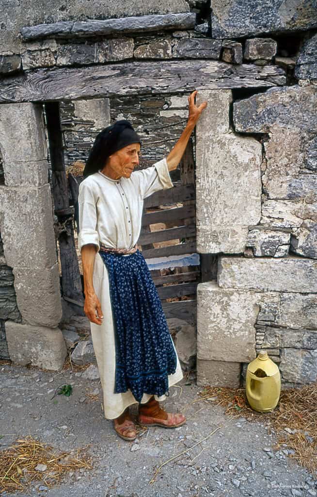 Elderly woman in traditional Olympos attire standing at a stone doorway