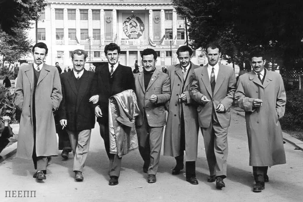 Group of Greek political refugees in Tashkent during the 1960s walking confidently down a city street in coats and suits, with a Soviet building decorated with a hammer and sickle emblem in the background.