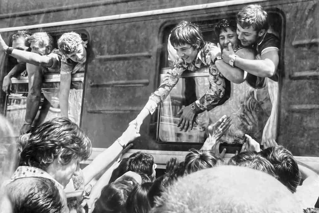 Young Greeks in the late 1970s leaning out of a train window in Tashkent, reaching out to say goodbye as they depart for Greece, surrounded by smiling friends and relatives waving from the platform.