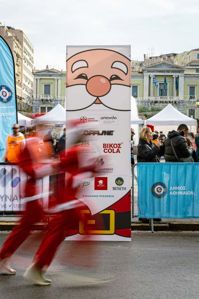 A runner dressed as Santa Claus passes in motion blur in front of Athens Santa Run signage during a festive city race