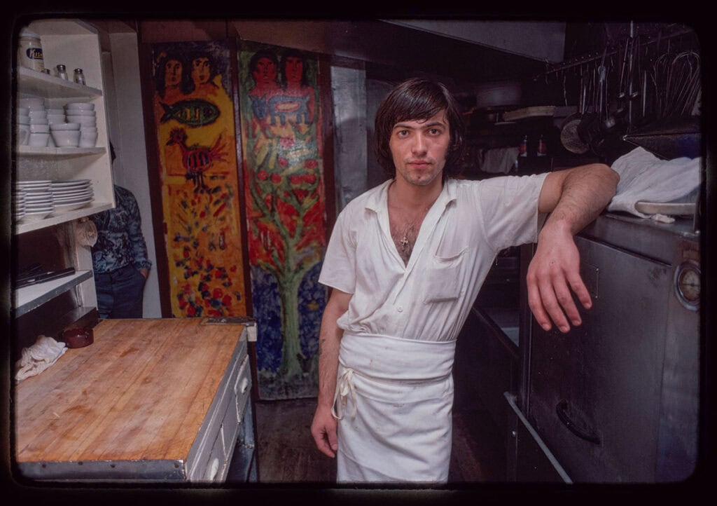 Restaurant worker standing in front of a painted mural inside a Greek restaurant
