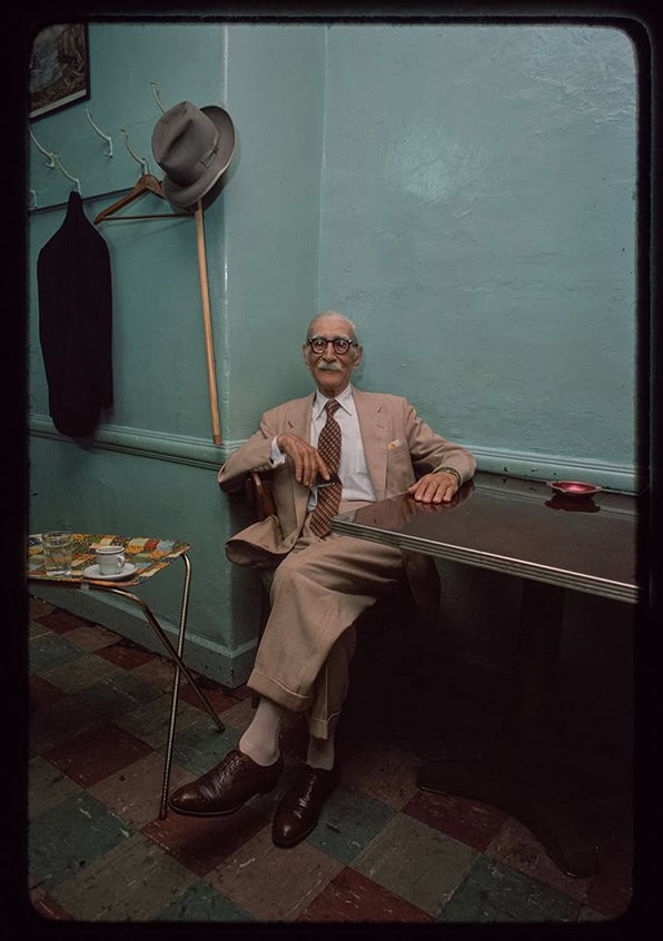 Elderly man in a suit seated alone at a small table inside a café