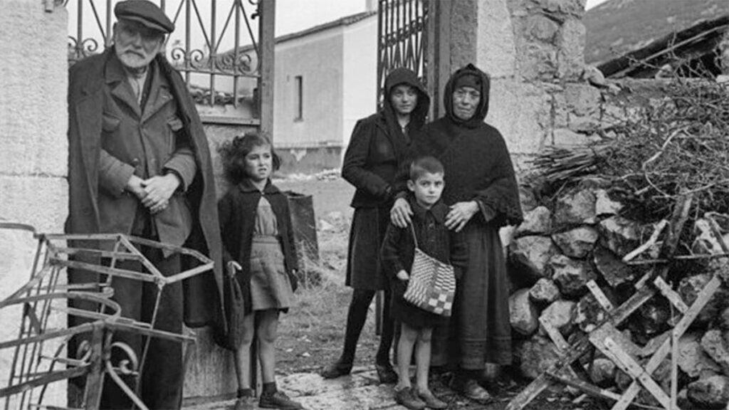 Black-and-white photograph of young Argyris Sfountouris standing with relatives outside a stone house in Distomo.
