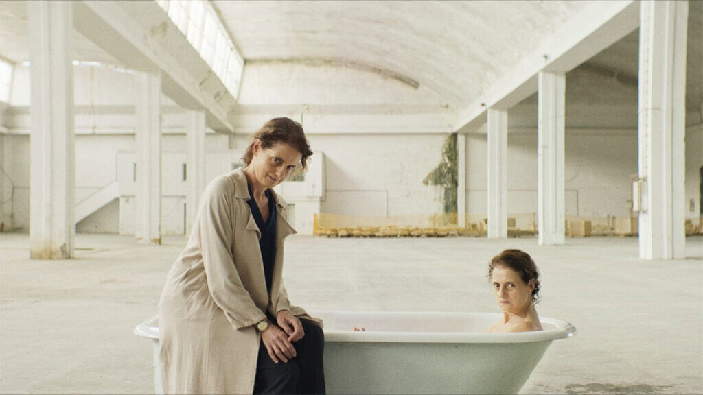 Two women in a bright industrial room, one seated beside a bathtub and one inside it, in a scene from Dreaming of Lions.