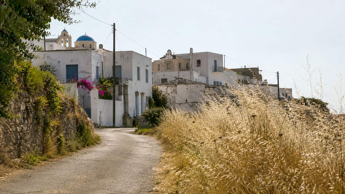 Dry grasses and weathered whitewashed houses along a quiet Greek hillside village road in mid-June