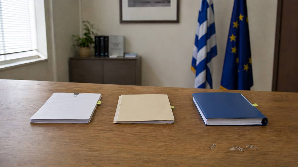 Document review table in an administrative office with Greek and European Union flags in the background.