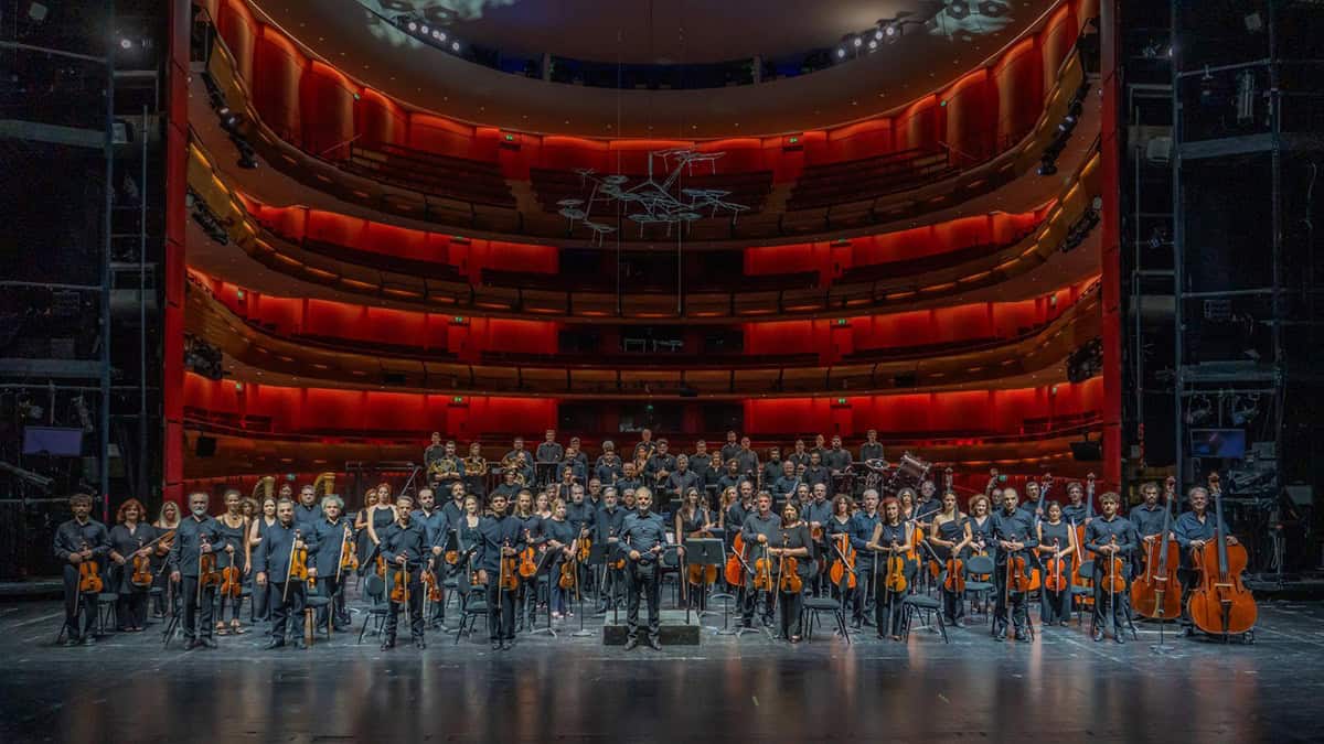 Members of the Greek National Opera Orchestra stand with their instruments onstage in a red-lit auditorium.