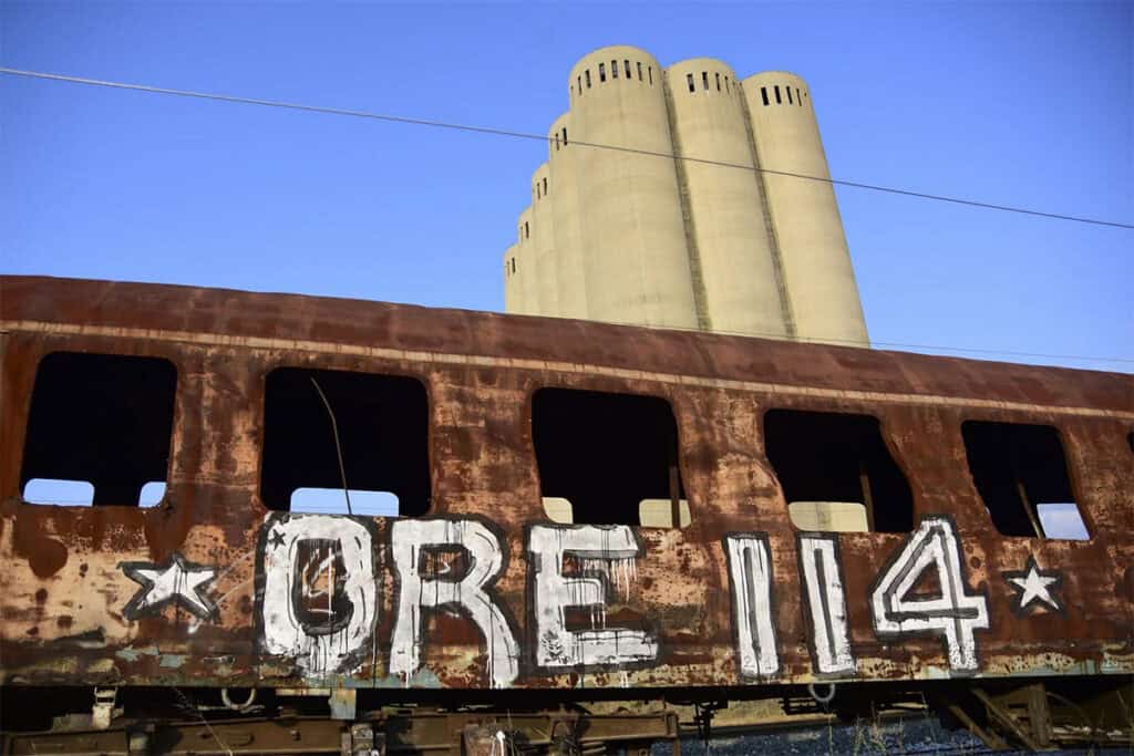 ORE 114 graffiti by Orestis Pangalos on a rusted railcar beneath industrial silos