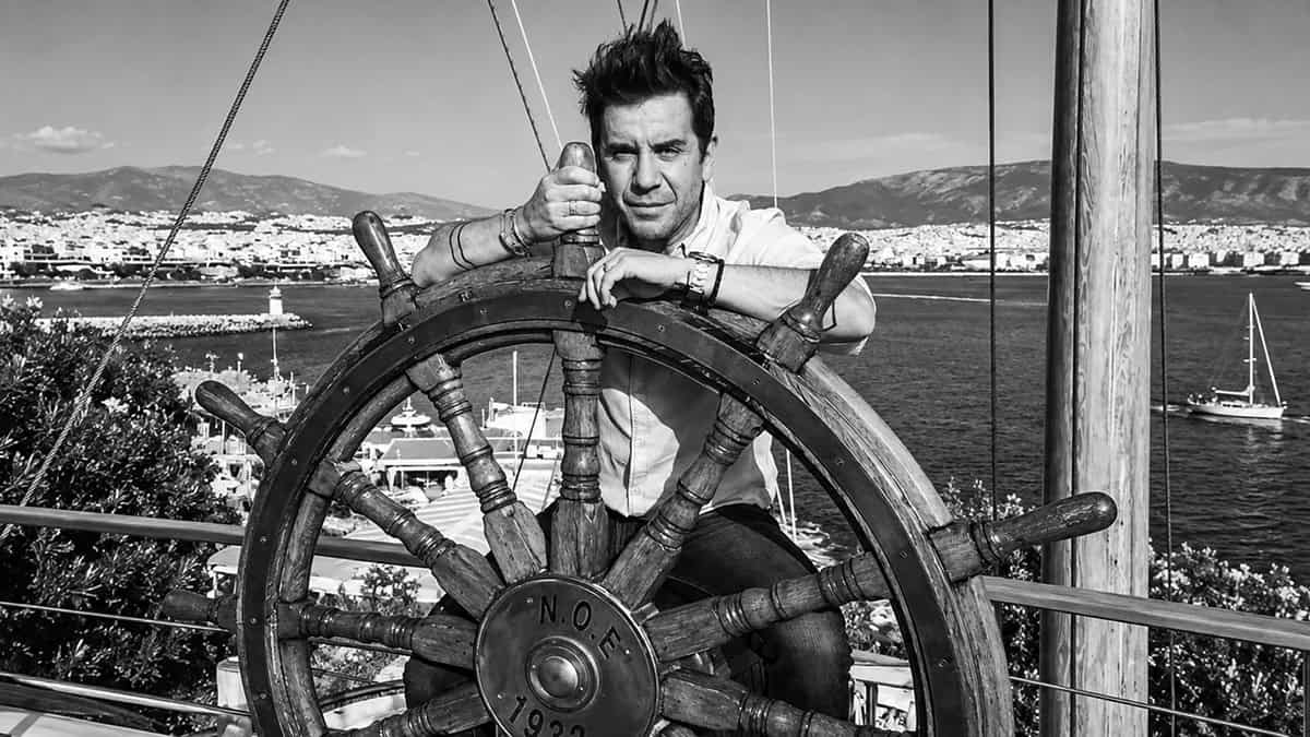 Greek singer Giorgos Mazonakis seated behind a large wooden ship’s wheel overlooking the Athens coastline