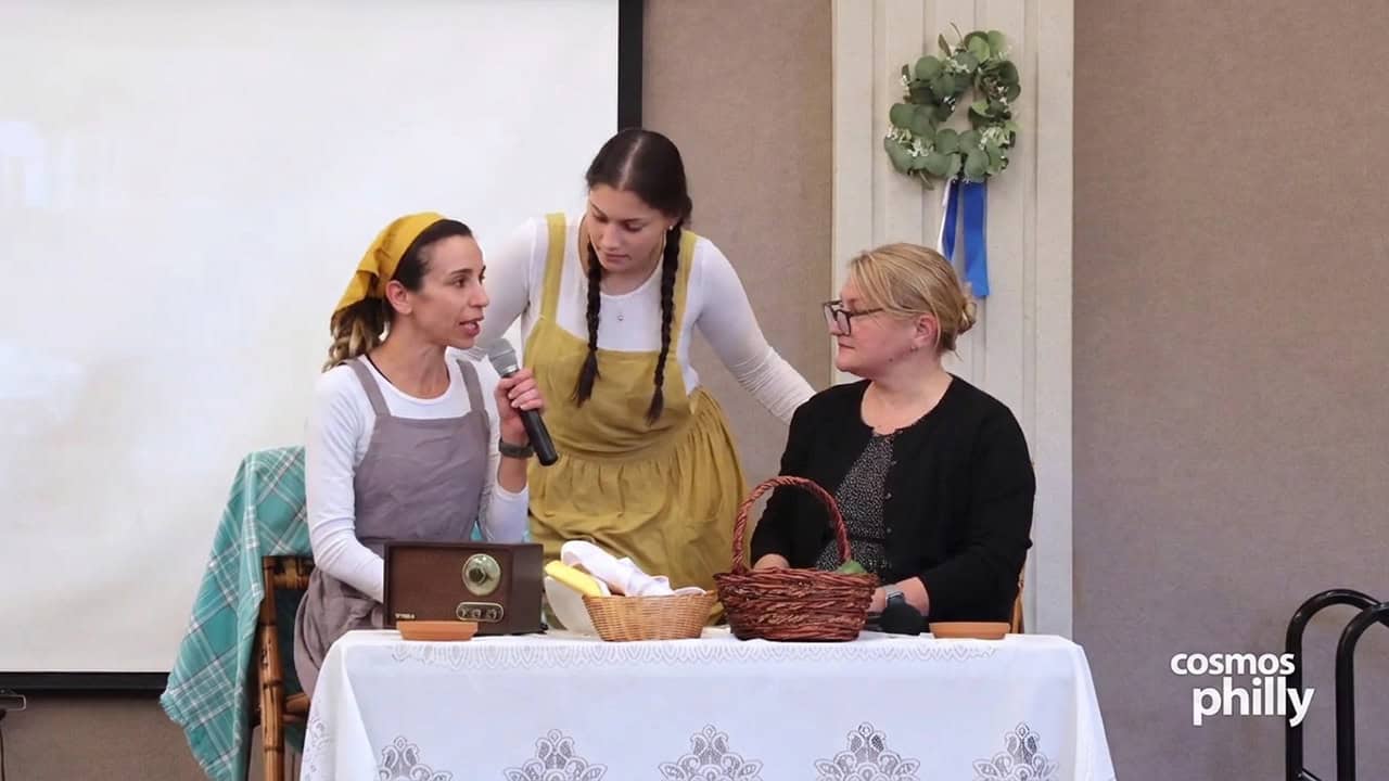 Three women perform a theatrical scene around a table with baskets and a vintage radio during Annunciation Greek School’s OXI Day celebration in Elkins Park.