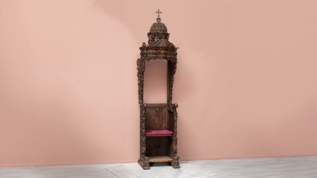 Tall carved wooden episcopal throne with an ornate canopy, cross, and red seat cushion.