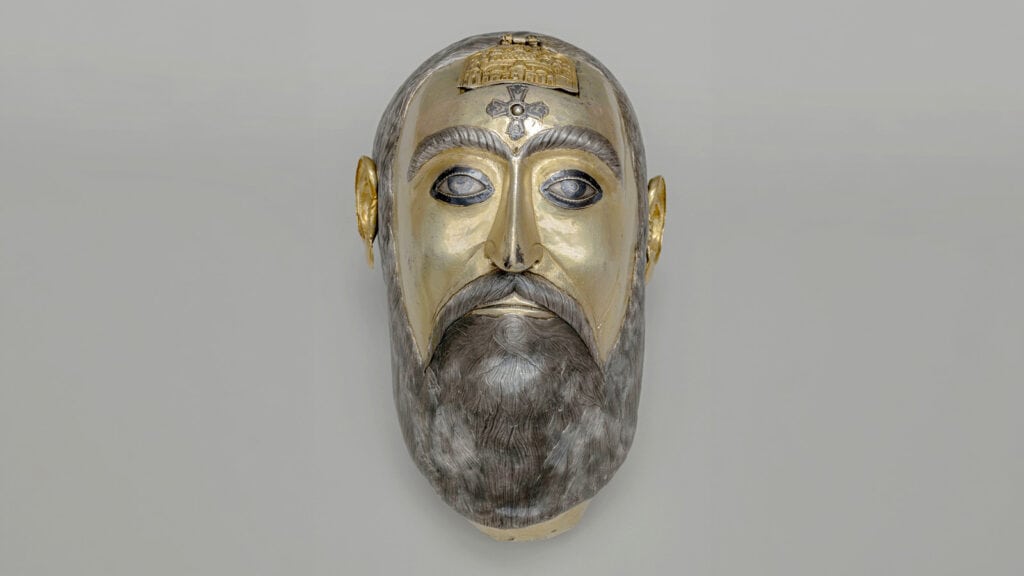 Head-shaped metal reliquary dated 1744, with a gold-colored face, silver beard, and cross on the forehead.