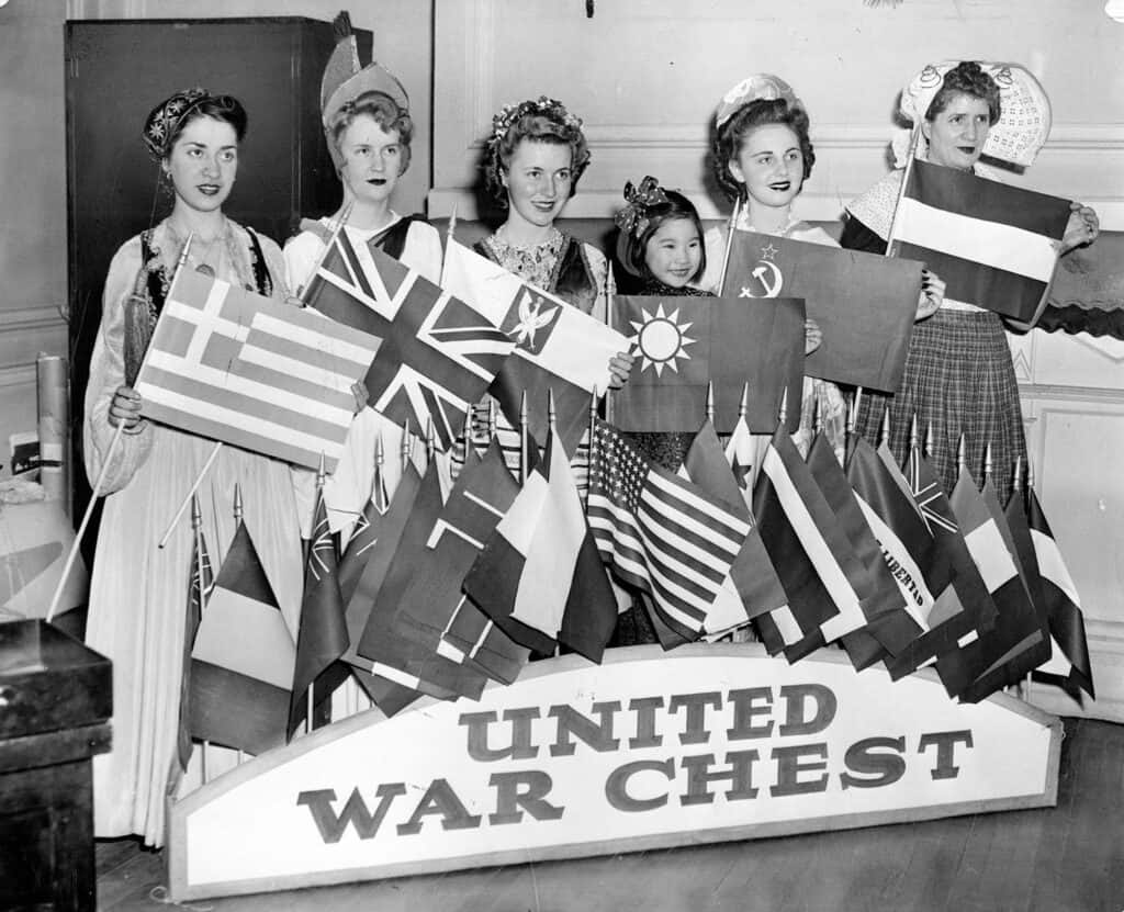 Six women dressed to represent different nations pose with Greek, British, Polish, Chinese, Soviet, and Dutch flags behind a large United War Chest sign.