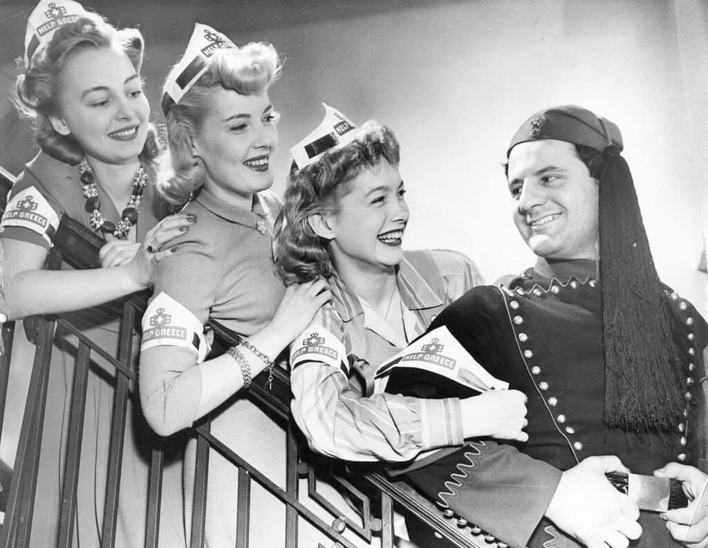 Three women wearing Help Greece armbands and paper caps pose with a smiling man in traditional Greek military dress at a wartime fundraising event.