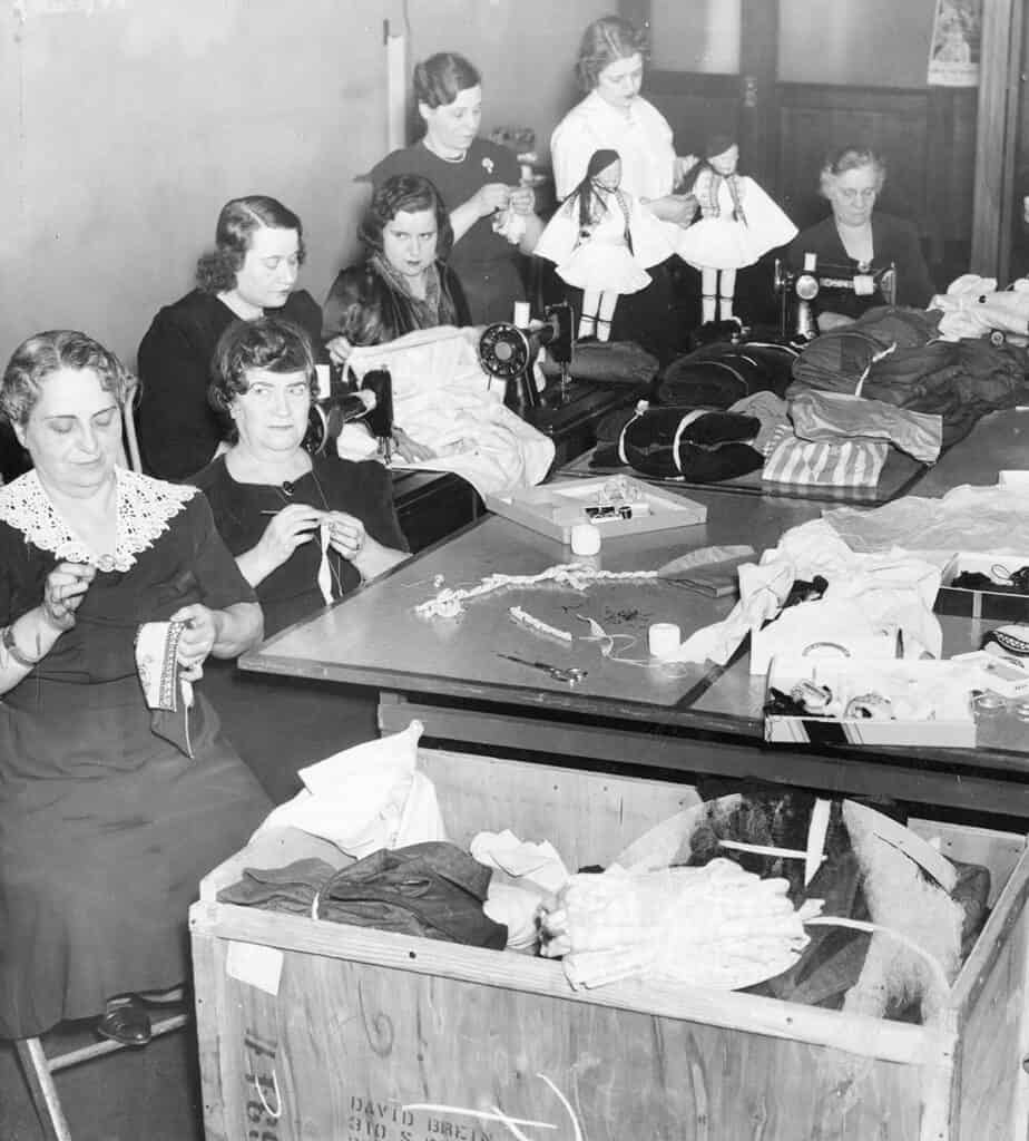 Seven women sew and knit garments around worktables covered with clothing, fabric, and sewing supplies, with two dolls dressed in Greek costumes behind them.